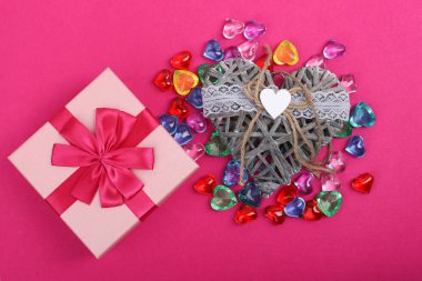 Kırmızı arka planda pembe bir kutu hediye ve yanında hasır dekoratif kalp. Hasır kalpli hediye kutusu, yanında renkli kalplerle çevrili. Kalp şeklinde mücevherler.