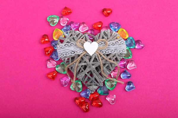 Decorative wicker heart surrounded with coloured hearts. Heart shaped gems on crimson background and wicker heart in center, top view. — Stock Photo, Image