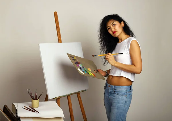 O artista no cavalete pinta um quadro. Menina atraente com um pincel tintas. Uma tela em branco para uma nova pintura e um artista com um pincel e tintas . — Fotografia de Stock