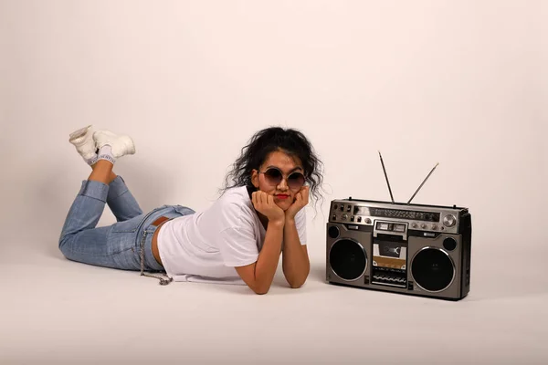 Chica feliz en una camiseta blanca y jeans miente y escucha música. Retro reproductor de cinta y chica. El estilo hip-hop es una chica joven que ama la música. Diferencia generacional y pasión por la música . Imagen De Stock