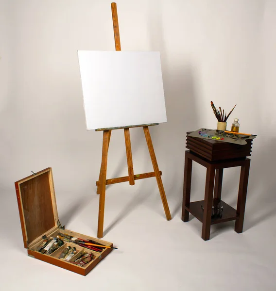 Oil painting. Blank canvas on Easel. Palette with paints and brushes. Squeeze out oil paints on a palette. Clean cloth with copy space. Set of brushes in a cup. Box with drawing tools