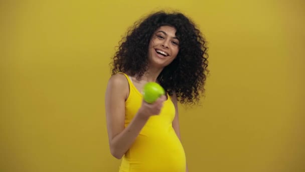 šťastný bi-rasové těhotné žena drží zralé jablko izolované na tmavě žluté