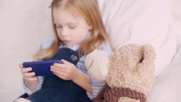 Yatakta Oyuncak Ayının Akıllı Telefonunu Kullanan Seçici Bir Çocuk — Stok video