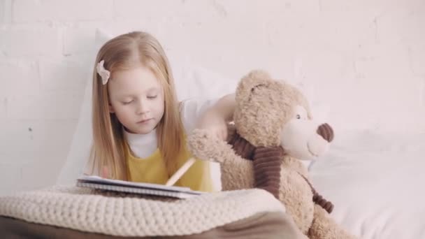 小孩抱着泰迪熊在床上画画 — 图库视频影像