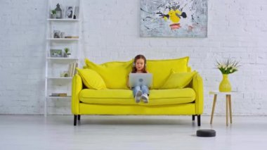 Kulaklıklı çocuk robot elektrik süpürgesinin yanında görüntülü sohbet yapıyor.