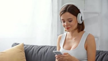 Akıllı telefonlu gülümseyen kız kanepede kulaklıkla müzik dinliyor.