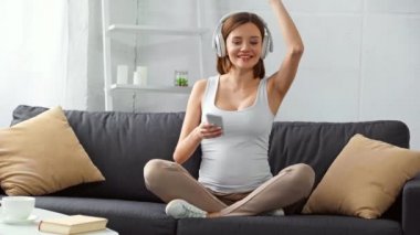 Kulaklıklı neşeli hamile kız elinde akıllı telefonuyla kanepede şarkı söylüyor. 