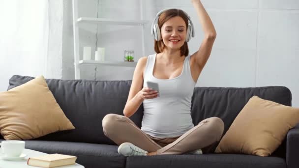 Chica Embarazada Alegre Auriculares Sosteniendo Teléfono Inteligente Cantando Sofá — Vídeo de stock