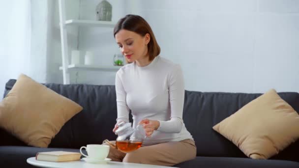 笑顔妊娠中の女性飲むお茶と読書本上のソファ — ストック動画