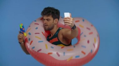 Yüzme ringinde oyuncak tabancalı bir adam mavi üzerine izole edilmiş akıllı telefonuyla selfie çekiyor.