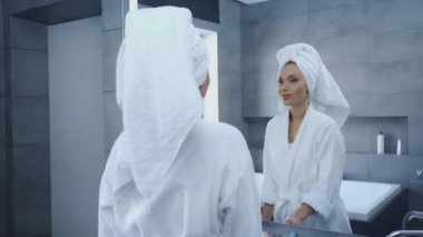 Bornozlu güzel bir kadın aynanın önünde makyaj fırçası kullanıyor.