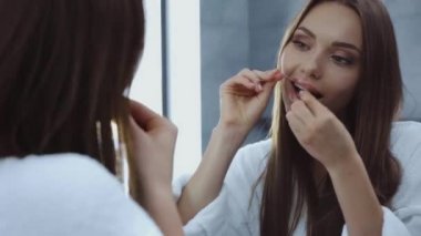 Güzel genç bir kadın dişlerini diş ipiyle temizliyor. Aynanın önünde.