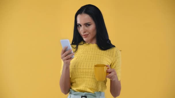Uśmiechnięta Dziewczyna Filiżanką Pomocą Smartfona Patrząc Aparat Odizolowany Żółty — Wideo stockowe
