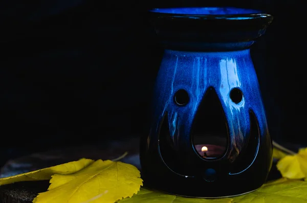 Lampada profumata in ceramica blu come Jack-o-lanterna con candela accesa in piedi su un ceppo con foglie autunnali gialle. Fondo nero. Concetto Halloween. Copia spazio — Foto Stock