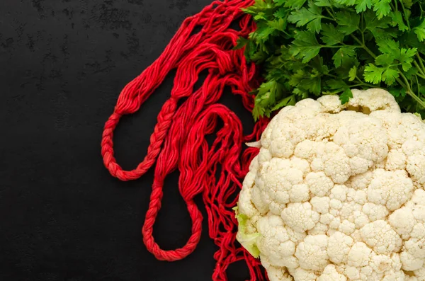 Red string bag, raw white cauliflower and fresh green parsley on black background. Eco friendly lifestyle. Zero waste concept. Flat lay, top view. Selective focus. Copy space