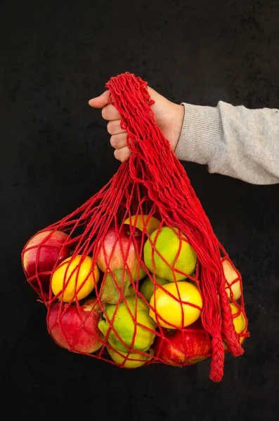 Main féminine tenant un sac à ficelle rouge avec des fruits sur fond noir. Concept zéro déchet. Style de vie écologique — Photo