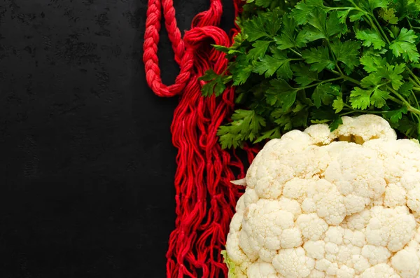 Red string bag, raw white cauliflower and fresh green parsley on black background. Eco friendly lifestyle. Zero waste concept. Flat lay, top view. Selective focus. Copy space