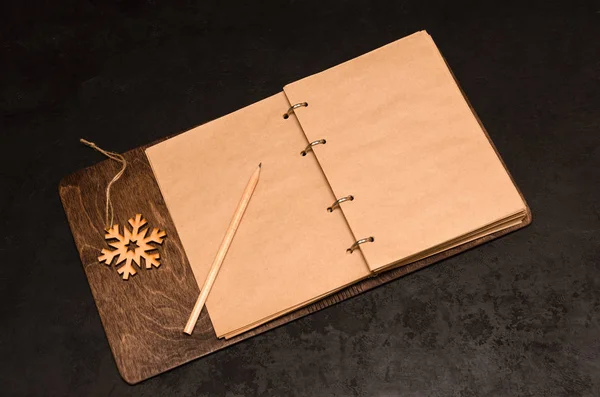 Zápisník Kraft s prázdnými listy v dřevěném obalu s prostorem pro text, tužku a dřevěnou sněhovou vločku na černém pozadí. Plán na příští rok. Na seznam. Vánoční pozadí. Mockup — Stock fotografie