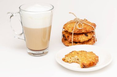 Şeffaf bir fincan sıcak latte ve beyaz arka planda katlanmış kuru üzümlü ev yapımı tam tahıllı yulaf ezmeli kurabiye. Kurabiyelerden biri ısırılıp koparılmış. Güne iyi başlangıç kavramı. Yakın çekim.