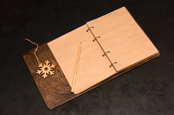 Σημειωματάριο με λευκά φύλλα Κραφτ σε ξύλινο κάλυμμα, μολύβι και ξύλινη νιφάδα χιονιού σε μαύρο φόντο. Έννοια των στόχων που τίθενται για το επόμενο έτος. Να κάνω λίστα. Χριστουγεννιάτικο φόντο. Κοπάδι — Φωτογραφία Αρχείου