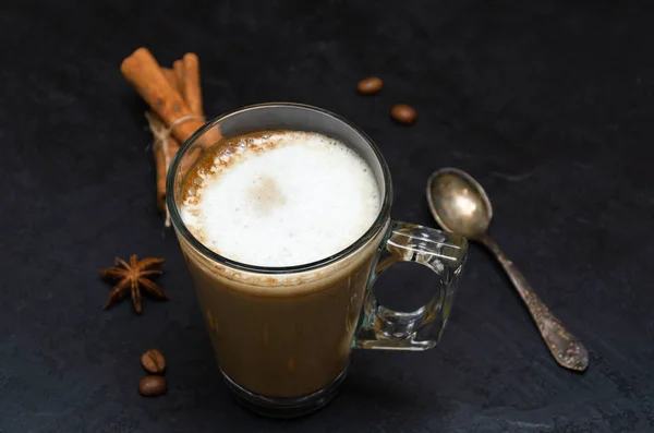 Διαφανής κούπα καπουτσίνο ή latte, σκορπισμένοι κόκκοι καφέ, κουτάλι, ξυλάκια κανέλας, αστεράκι γλυκάνισου σε μαύρο φόντο. Ώρα για καφέ και χαλάρωση. Θέα από ψηλά. Κοντινό πλάνο — Φωτογραφία Αρχείου