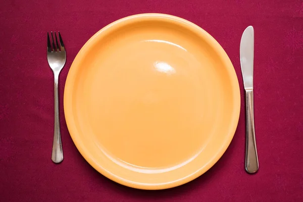 Empty yellow plate, fork and knife on maroon tablecloth. Elegant table setting. Top view, closeup — Stock Photo, Image
