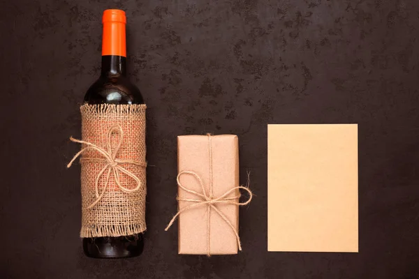 Bottle of red wine, gift box wrapped in kraft paper and blank card for your greetings on black background. Zero waste holidays and eco-friendly gift wrapping concept. Flat lay, top view