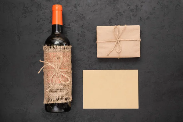 Red wine bottle, gift box wrapped in kraft paper and blank card for your message on black background. Zero waste holidays and eco friendly gift wrapping concept. Flat lay, top view