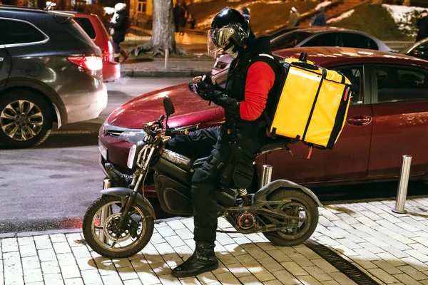 Kiev, Ucrânia - 24 de janeiro de 2020. Yaroslaviv Val. Correio de entrega de alimentos com grande mochila amarela sentada em scooter e tomando ordem via smartphone na rua da cidade à noite. Serviço de entrega rápida — Fotografia de Stock