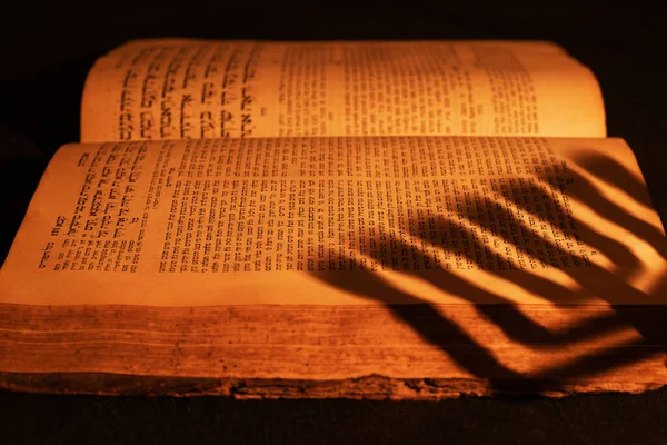 Stara hebrajska Biblia w świetle płonącej świecy na ciemnym tle. Cień z menory na otwartym żydowskim modlitewniku w ciemności. Zbliżenie — Zdjęcie stockowe