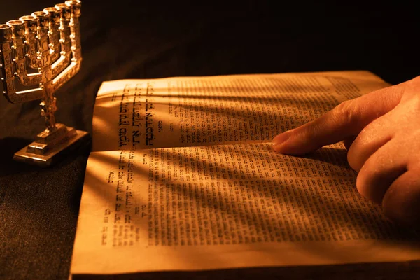 Old Hebrew Bible and silver menorah in light of burning candle on dark background. Shadow from menorah on open pages of Jewish prayer book in the darkness. Torah reading. Close-up — Stock Photo, Image