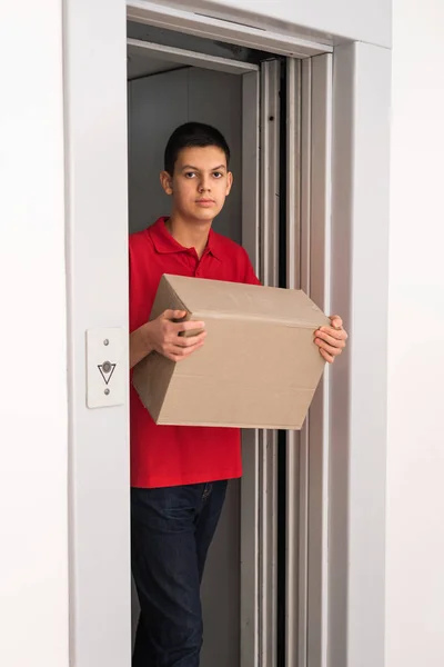 Young friendly delivery courier dressed in red polo shirt leaving the elevator holding cardboard box with order. Delivery service and logistic concept. Part-time job for teenagers and students
