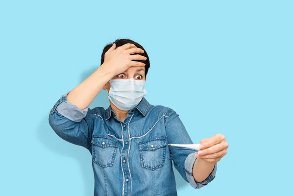 Astonished sick woman in medical mask looking with expression at thermometer in hand on light blue background. Middle-aged brunette surprised of high temperature. Coronavirus and flu epidemic concept