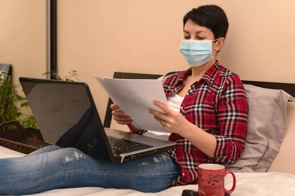 Tıbbi koruyucu maskeli bir kadın evde dizüstü bilgisayarla karantina ya da tecrit sırasında çalışıyor. Coronavirüs salgını, covid-19 salgını. Uzaklık çalışması, e-öğrenme, çevrimiçi eğitim