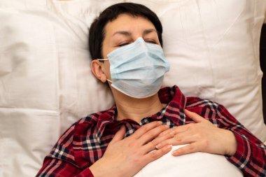 Hasta, maske takmış, göğüs ağrısı çeken ve nefes nefese kalmış genç bir kadın. Koronavirüs salgını sırasında evde kalan kısa saçlı esmer ve covid-19 veya zatürree belirtileri var.