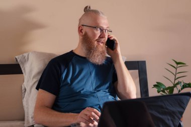 Evde çalışan orta yaşlı iş adamı dizüstü bilgisayarla çalışırken cep telefonuyla konuşuyor. Gülümseyen kızıl sakallı adam iş sorunlarını uzaktan çözer. Mesafe çalışması ve serbest çalışma kavramı