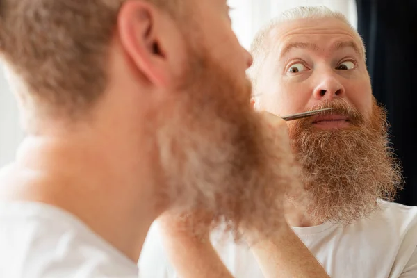 Attractive middle-aged man cutting his moustache and beard by himself in front of mirror at home. Beard care during quarantine. Coronavirus outbreak, lockdown, staying home on self-isolation. Closeup