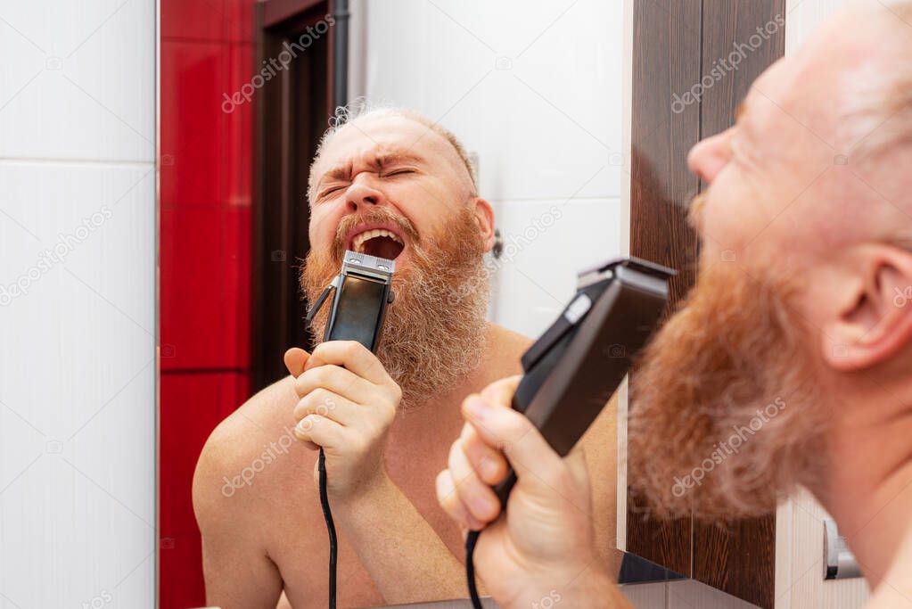 Handsome cheerful bearded man singing into hair trimmer instead of microphone in front of mirror in bathroom at home. Positive happy man joking and portraying a singer. Closeup
