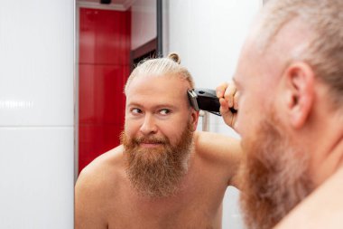 Yakışıklı sakallı adam evde aynanın önünde banyoda saç makasıyla kendi saçını kesiyor. Karantina sırasında saç bakımı. Yakın çekim.