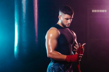 Genç ve kaslı bir adam ellerini boks elleriyle bantlıyor. Dövüş, spor ve boks temaları.