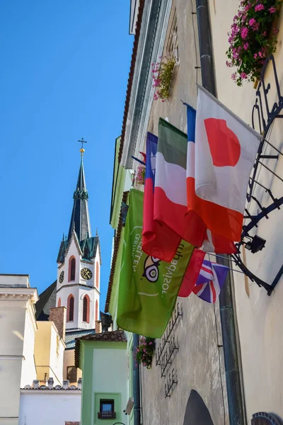 Český Krumlov, Česká republika - Oktober 11, 2018: Katolická kostelní věž s hodinami a vlajkami zemí na zdi — Stock fotografie