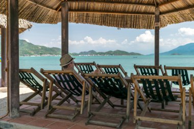Young man with hat sits in a chair by sea under a canopy and look at the sea on vacation
