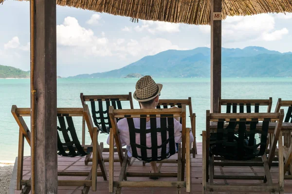 Young man with hat sits in chair on beach by sea under canopy and look at sea on vacation