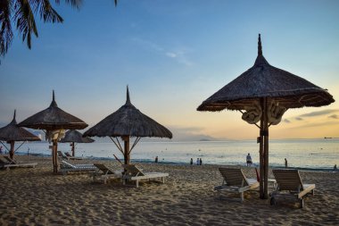 Şafak vakti kumlu bir sahilde saman şemsiyeler ve deniz kenarında ahşap şezlonglar.