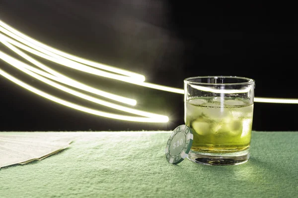 Glass misted glass with a drink and ice on the green table, the glare of light, drawing light. Poker chip next, dollars in the background