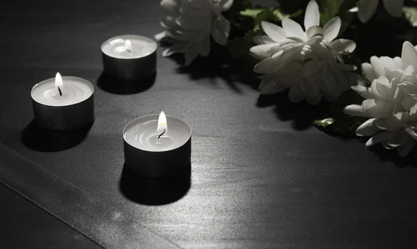 Funeral symbol. A bouquet of white flowers and burning candles, black background, free space for text. Satin ribbon in the corner.