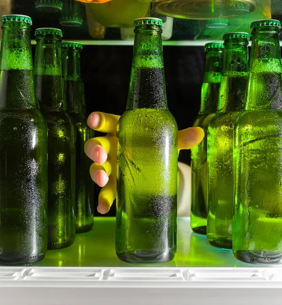 The hand reaches for the beer bottle. Green bottles of beer and drops of condensation are on a shelf in the refrigerator.