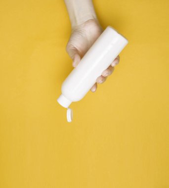 Beyaz plastik şampuan şişesi ya da başka kozmetik ürünleri olan bir kadın eli. Sarı arkaplan, kozmetik ürünler.