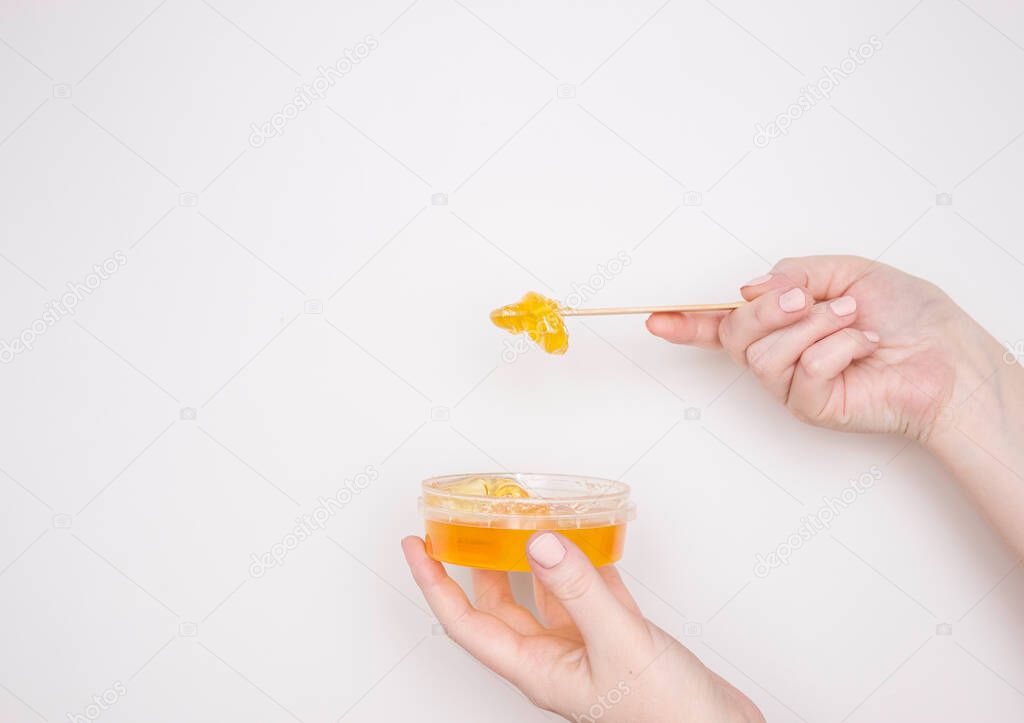 A woman's hand with a wooden spatula and sugar paste for depilation and a jar. Light background, copyspace.