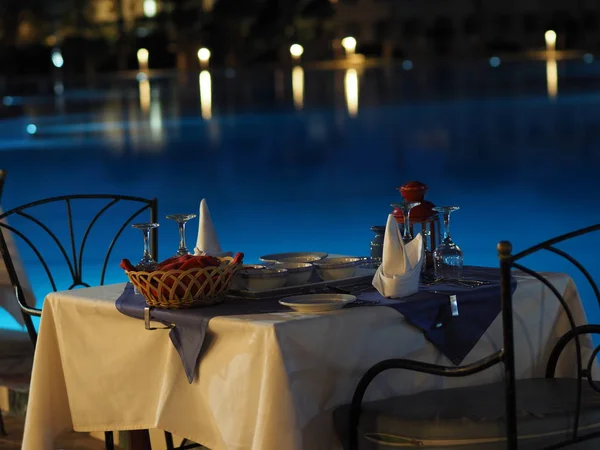 served tables at a restaurant with no people on the street preparing to welcome visitors against a backlit evening pool